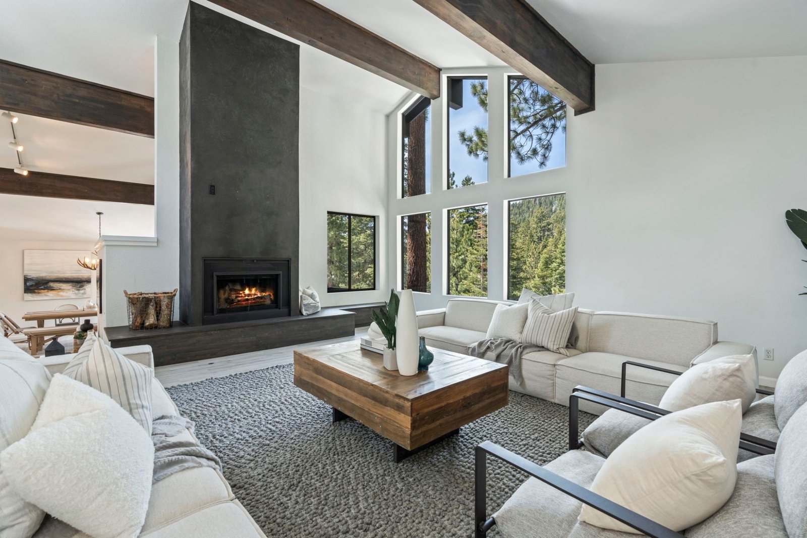 Bright vaulted living room in an Incline Village home with exposed dark wood beams, a floor-to-ceiling charcoal fireplace, and tall windows looking out to tall pines