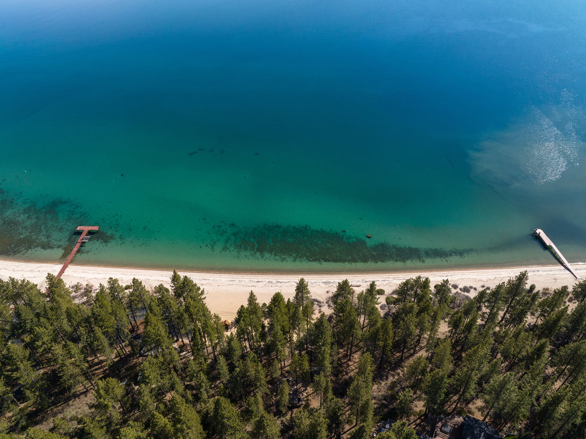 Aerial view of a pine-lined Lake Tahoe lakefront with a white sand beach, two private piers, and clear turquoise water