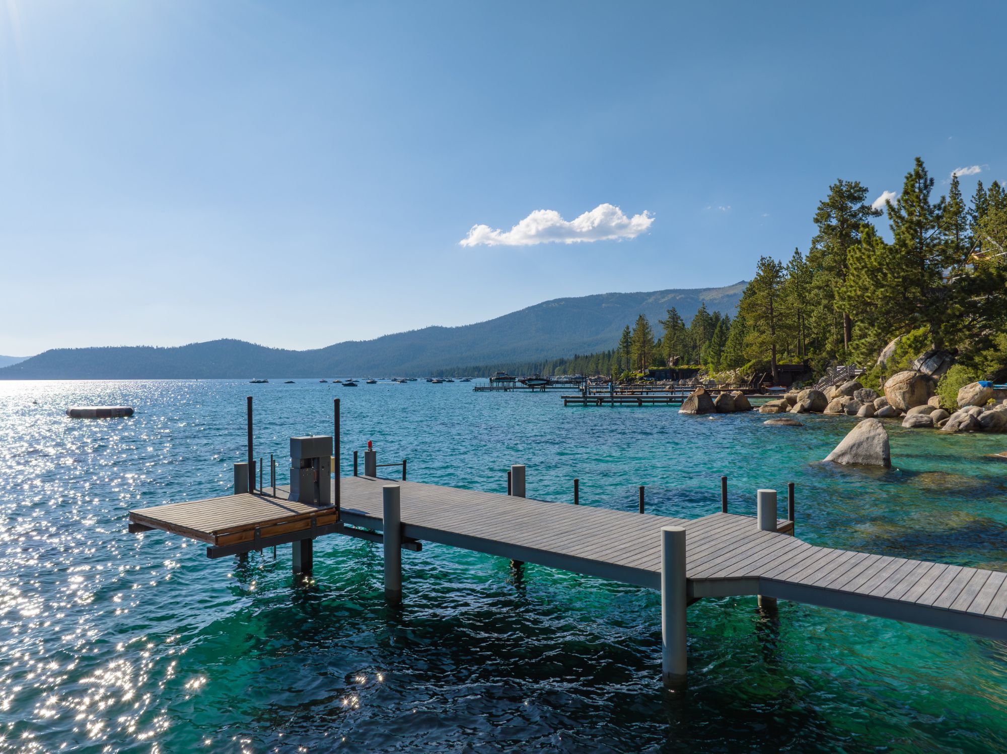 A private pier with a boat lift over clear turquoise Lake Tahoe water, beside a granite-boulder shoreline lined with pines, with moored boats and forested mountains beyond